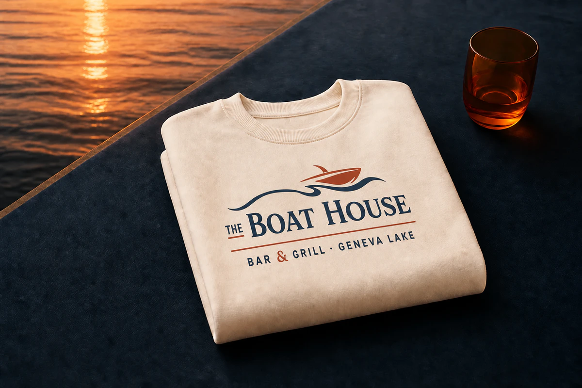 The full two-color Boat House lockup screen-printed into a warm-cream crewneck beside Buttons Bay at sunset.